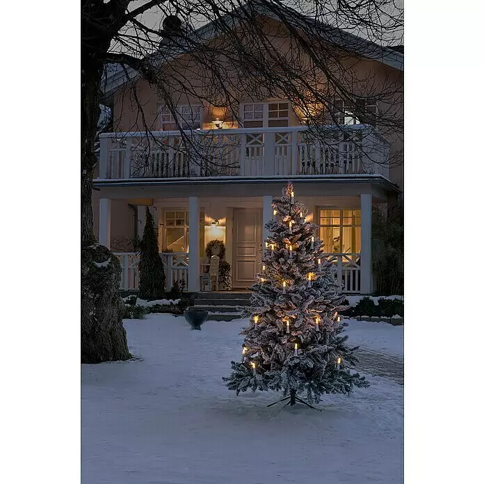 Konstsmide LED-Weihnachtskerzen - 21,8 mm, Weiß, Lichtfarbe: Bernstein, 25 Stk., IP44 - Beflockter Outdoor-Weihnachtsbaum mit warmweißen Kerzenlichtern steht im Schnee und schafft eine festliche Atmosphäre vor einem gemütlichen Haus.