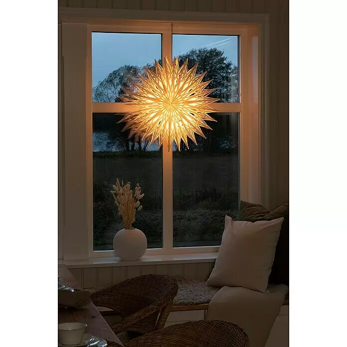 Konstsmide LED-Stern - 60 cm, Weiß, Netzbetrieben - Eine leuchtende, weiße Papiersternlampe hängt im Fenster, spendet warmes Licht nach drinnen und ist von außen sichtbar vor dunkler Landschaft.