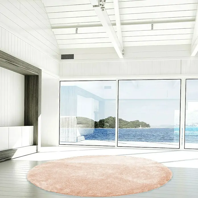 Kayoom Hochflorteppich Diamond - Puderrosa, Durchmesser: 160 cm, 100 % Polyester - Ein weicher, runder Teppich in zartem Rosa liegt in einem hellen Raum mit Blick auf das Meer. Er sorgt für Wärme und Komfort.