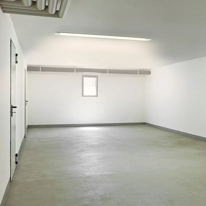 Ritter Leuchten LED-Feuchtraumlichtleiste Slim Pro - L x B x H: 146 x 4,3 x 3,3 cm, Lichtfarbe: Neutralweiß, 45 W, IP65 - Leerer Raum mit Betonboden, weißen Wänden, Fenster und Tür. Eine lange Leuchtstoffröhre sorgt für die Beleuchtung des Zimmers.