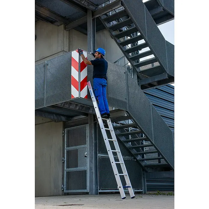 Krause Stabilo Mehrzweckleiter +S - Anzahl Tritte: 30 Stk., Arbeitshöhe: 7,5 m, Aluminium - Mann auf einer Aluminium-Leiter von KRAUSE, der an einem Gebäude arbeitet, mit Metalltreppe im Hintergrund.