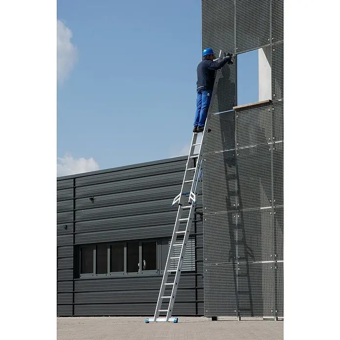 Krause Stabilo Schiebeleiter +S - Anzahl Tritte: 24 Stk., Arbeitshöhe: 6,7 m, Aluminium - Ein Mann auf einer KRAUSE Leiter vor einem Gebäude mit Metallgitterfassade bei Arbeiten an einem Fenster.