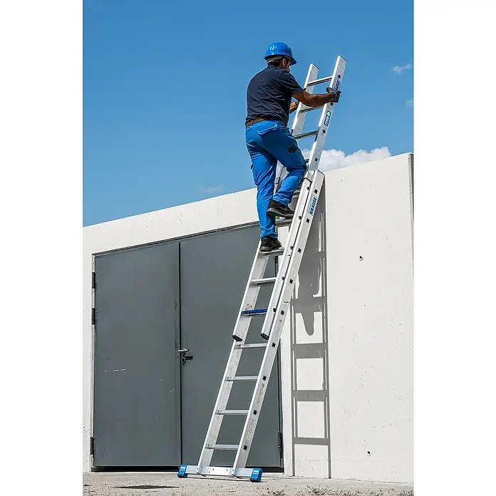 Krause Stabilo Schiebeleiter +S - Anzahl Tritte: 18 Stk., Arbeitshöhe: 5,15 m, Aluminium - Ein Handwerker mit Helm steigt eine Krause Leiter hoch, die an einer weißen Wand mit grauen Türen steht.