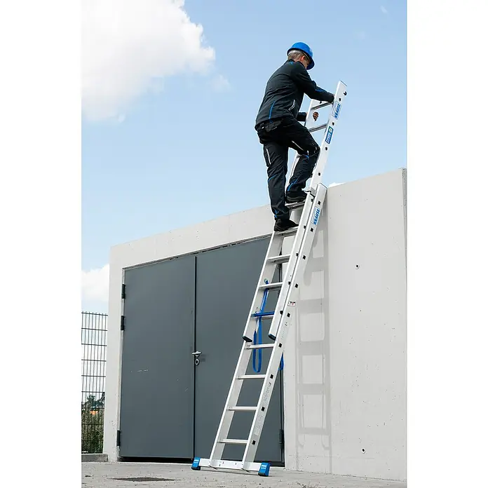 Krause Stabilo Mehrzweckleiter +S - Anzahl Tritte: 18 Stk., Arbeitshöhe: 5,15 m, Aluminium - Ein Mann mit Helm steigt eine Krause-Leiter an einer Betonwand hoch, vor einem grauen Gebäude mit bewölktem Himmel.