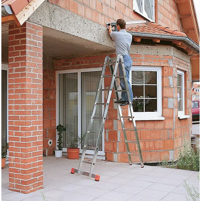 Krause Monto Doppelleiter TriMatic - Anzahl Tritte: 16 Stk., Arbeitshöhe: 5,5 m, Aluminium - Mann auf einer silbernen Mehrzweckleiter, der ein Werkzeug benutzt, vor einer roten Backsteinwand.