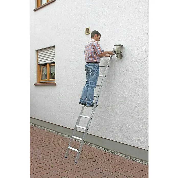 Krause Stabilo Anlegeleiter + S - Anzahl Tritte: 10 Stk., Arbeitshöhe: 3,6 m, Aluminium - Mann auf einer KRAUSE-Leiter, der eine Außenleuchte an einer weißen Wand repariert, mit Backsteinen als Bodenbelag.
