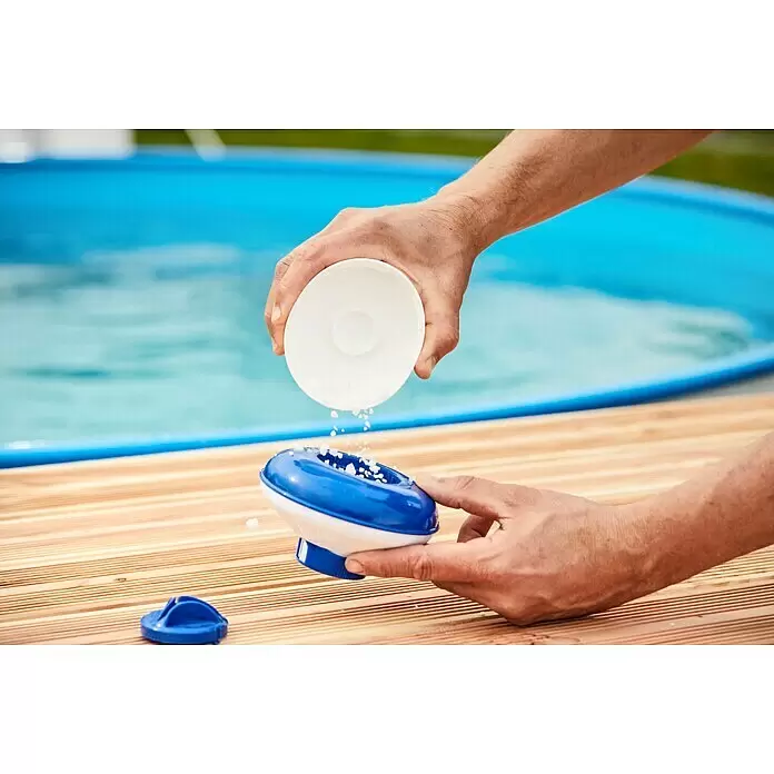 Malibu Schwimmdosierer - Geeignet für: Tabs - Hände füllen einen blau-weißen Pool-Chemikaliendosierer mit Granulat, neben einem Pool.