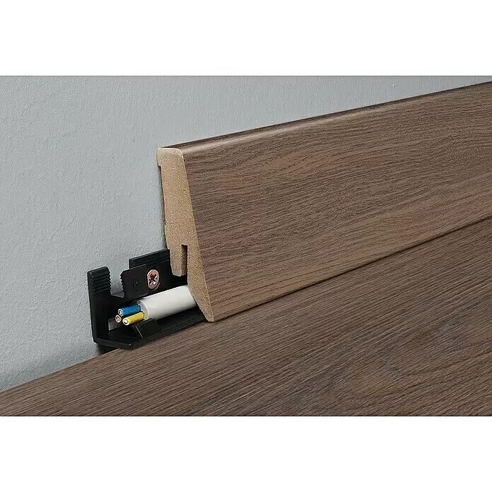 MyStyle Sockelleiste K58 C Bourbon Hill Oak - 260 cm x 18 mm x 58 mm, MDF - Braune Holzoptik-Sockelleiste mit integriertem offenem Kabelkanal, der ein weißes Kabel zeigt, an einer Wand montiert.
