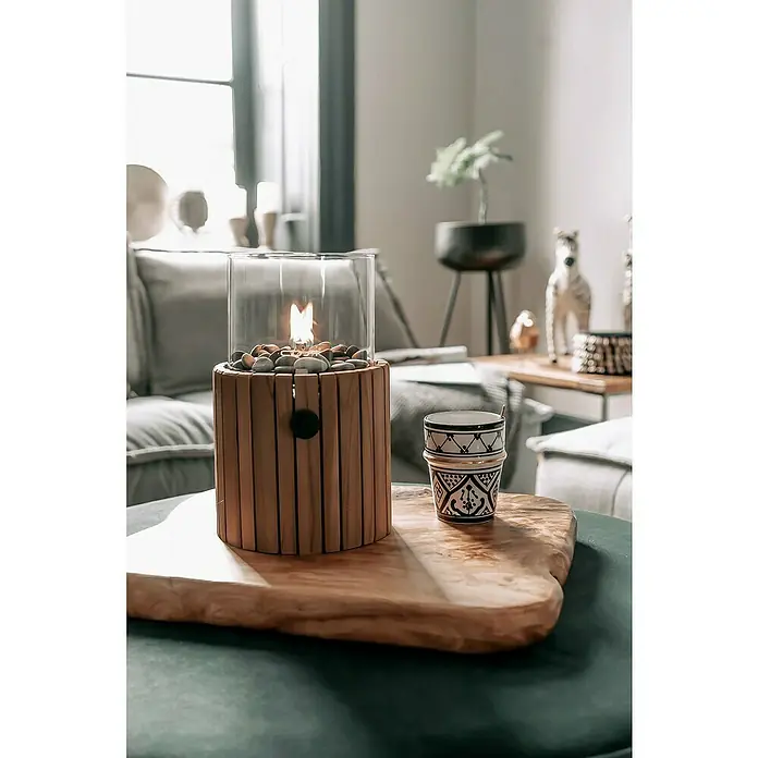Gaslaterne Cosiscoop Timber - Holz, Ø x H: 18,5 x 30 cm, Rund - Ein Tischfeuer mit Holzsockel und Glaszylinder, dazu eine dekorative Tasse auf einem Holzbrett.