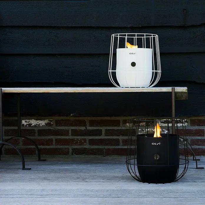 Gaslaterne Cosiscoop Basket - Black, Ø x H: 31 x 30,6 cm - Zwei moderne Tischfeuerstellen, eine weiße auf einer Bank und eine schwarze am Boden, beide mit dekorativen Metallkäfigen umgeben.