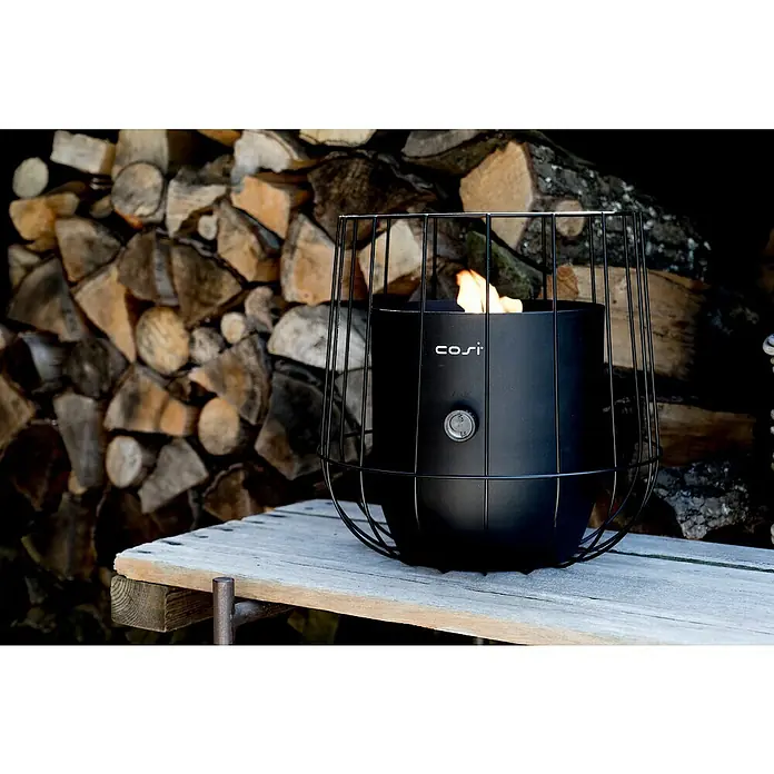Gaslaterne Cosiscoop Basket - Black, Ø x H: 31 x 30,6 cm - Nahaufnahme eines Cosi Feuertisches mit Flamme, umgeben von einem schwarzen Käfig, auf einer Holzoberfläche vor einem Holzstapel.