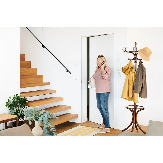 Nuki Elektrischer Türöffner Opener - Passend für: Nuki Smart Lock - Eine Frau telefoniert im Flur neben einer Holztreppe. Der Flur verfügt über eine Garderobe und einen Teppich mit gelbem Muster.