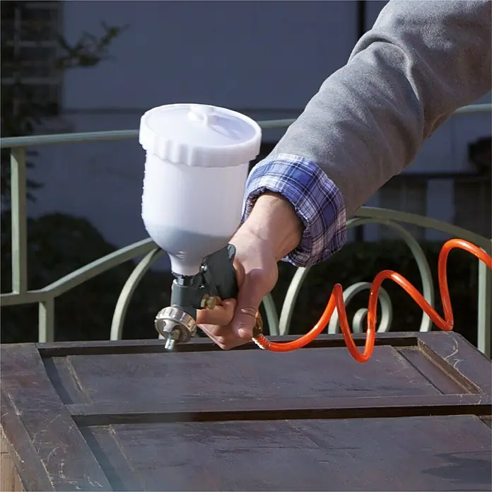 Herkules Pro-Line Flüsterkompressor TB 50V - 1.100 W, 50 l - Nahaufnahme einer Person, die mit einer Farbspritzpistole ein Möbelstück besprüht, wobei der Fokus auf der Spritzpistole und dem Möbelstück liegt.