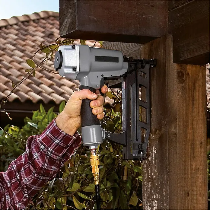 Herkules Pro-Line Flüsterkompressor TB 50V - 1.100 W, 50 l - Der Druckluftnagler wird an einem Holzpfosten befestigt, im Hintergrund sind Ziegel und Pflanzen zu sehen.