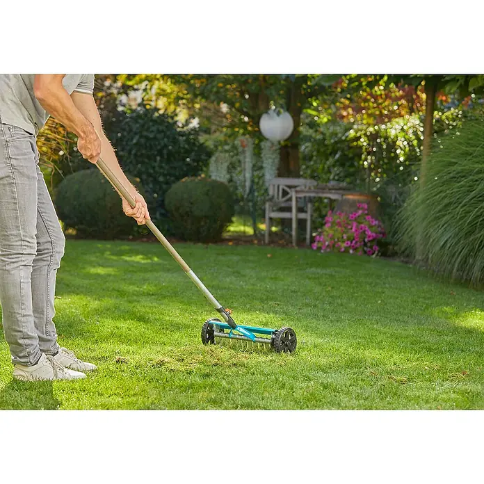 Gardena Combisystem Vertikutierboy - Arbeitsbreite: 32 cm, Geeignet für: Moos- und Rasenfilzentfernung - Eine Person benutzt einen blauen Rasenlüfter mit Rädern auf einem grünen Rasen. Das Gerät dient zur Rasenpflege im Garten.