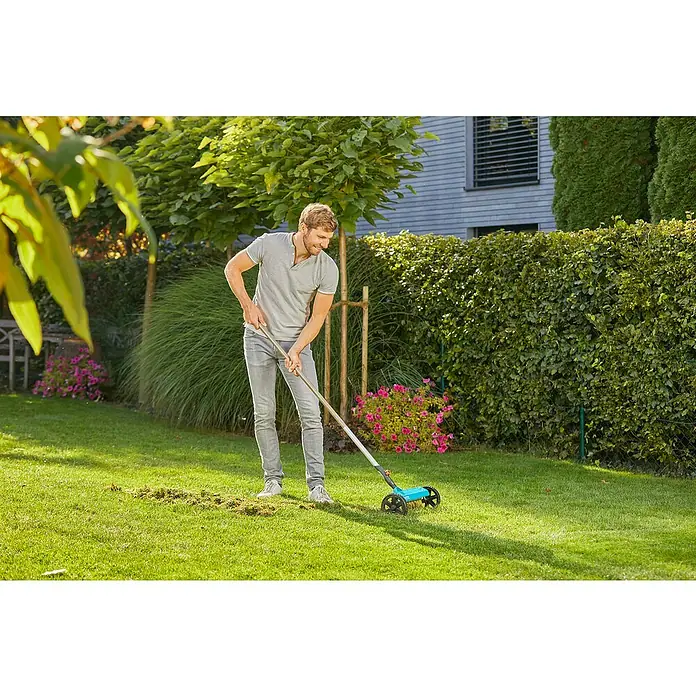 Gardena Combisystem Vertikutierboy - Arbeitsbreite: 32 cm, Geeignet für: Moos- und Rasenfilzentfernung - Ein Mann benutzt einen Rasenkehrer, um Laub auf einem grünen Rasen zu entfernen, während er im Garten steht.