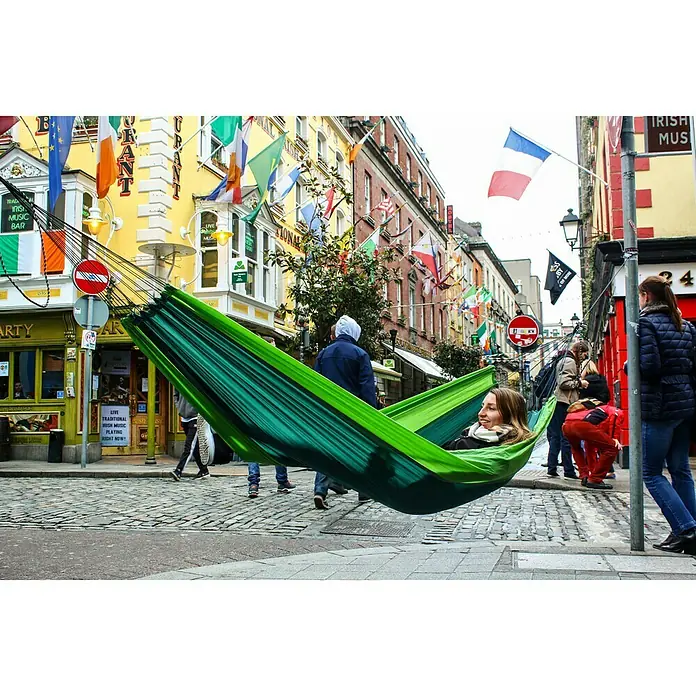 Amazonas Hängematte Silk Traveller forest - L x B: 220 x 140 cm, Grün - Eine Frau entspannt in einer grünen Hängematte, die zwischen Pfosten in einer belebten Stadt gespannt ist. Im Hintergrund sind Flaggen und Passanten zu sehen.