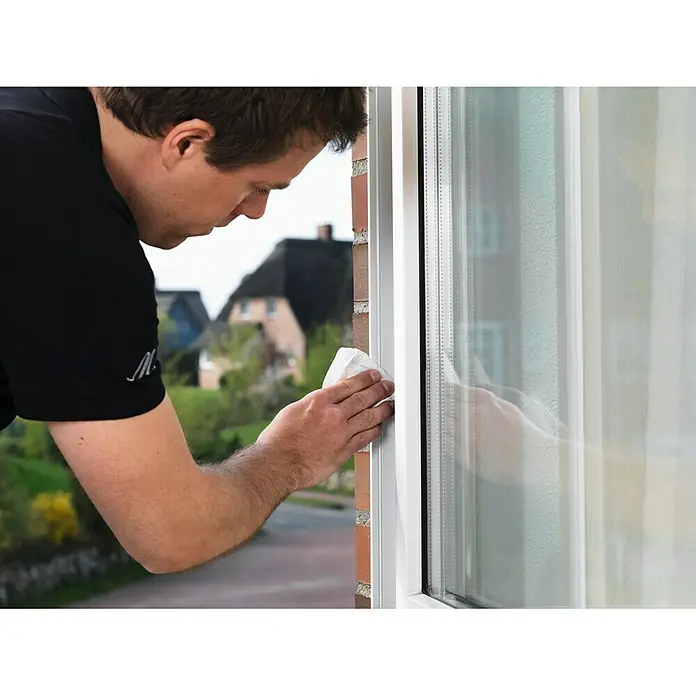 Tesa INSULATION Dichtungsband GUMMI L - Braun, 6 m x 9 mm x 5,5 mm, Geeignet für: Spaltenbreiten 2 - 5 mm - Mann reinigt Fensterrahmen mit einem Tuch. Das Fenster ist weiß und befindet sich in einem Haus mit Ziegelmauerwerk.