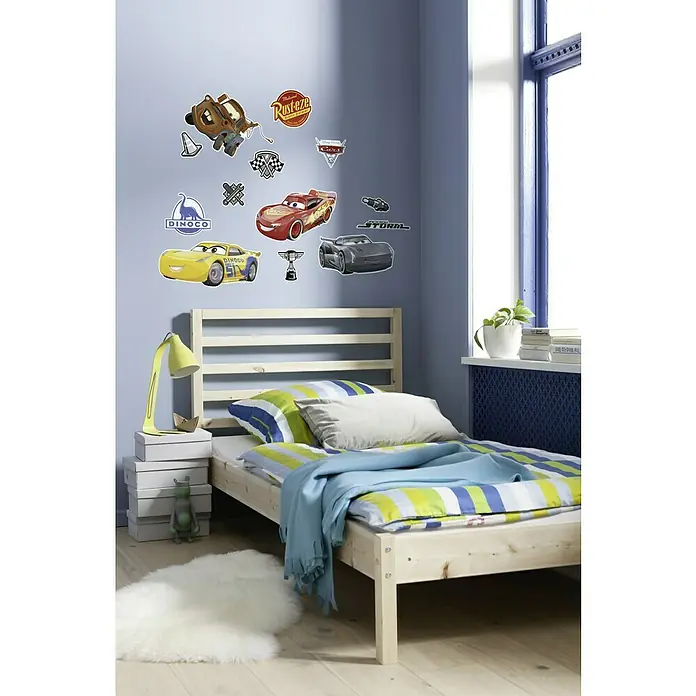 Komar Dekosticker Cars 3 - 70 x 50 cm, 13 -tlg. - Ein helles Kinderzimmer mit einem Holzbett, Cars Wandstickern und einer gelben Lampe auf einem Stapel von Kisten.