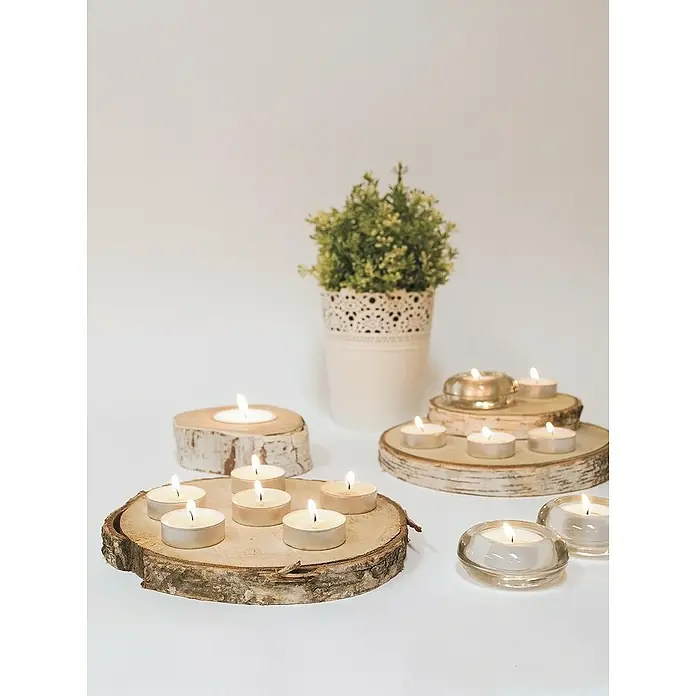 Ceras Roura Vela de té - Ø x Al: 38 mm x 16 cm, Blanco, Tiempo de combustión aprox.: 4 h, 50 ud. - Una foto de cerca de luces de té sobre rodajas de madera, junto a una pequeña maceta con una planta verde sobre una mesa blanca.