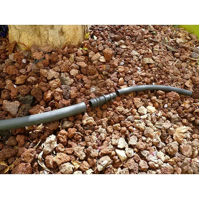 Manguito de unión reductor goteo 18 x 16 mm - 18 mm - 16 mm, 3 ud. - Primer plano de una tubería flexible gris con un conector negro sobre roca de lava roja. Práctico sistema de riego para el jardín.