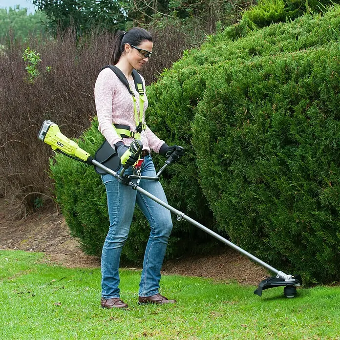 Ryobi ONE+ Akku-Rasentrimmer RBC 18X20B4F - 18 V, 4 Ah, Li-Ionen, 1 Akku, Schnittbreite Messer: 20 cm, Schnittbreite Faden: 30 cm - Eine Frau benutzt einen Ryobi Akku-Rasentrimmer mit Tragegurt, um Gras unter einem Busch zu schneiden.