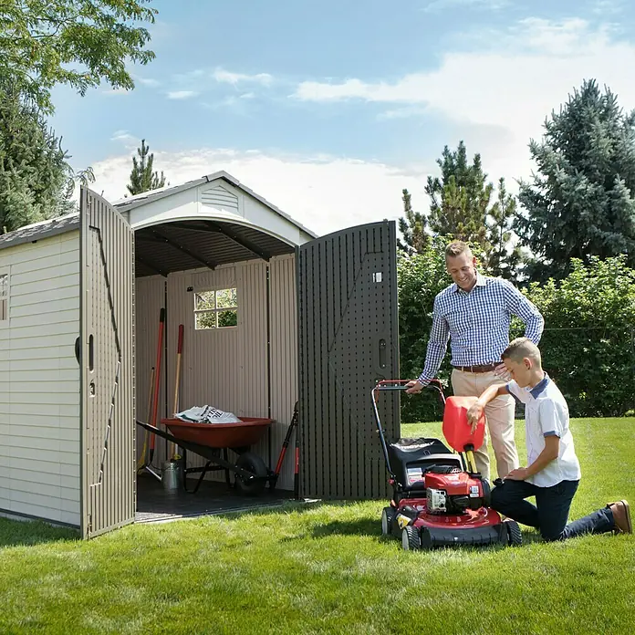 Gerätehaus Phoenix - Außenbreite inkl. Dachüberstand: 206 cm, Außentiefe inkl. Dachüberstand: 280 cm, Kunststoff, Lichtgrau/Anthrazit - Ein Vater und sein Sohn betanken einen Rasenmäher vor einem geräumigen Gartenschuppen mit geöffneten Türen, der viel Stauraum bietet.