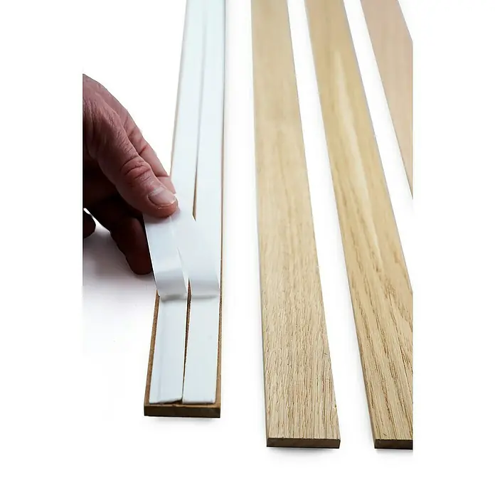 Panel de pared decorativo Sticks - Roble natural, 250 cm x 3,45 cm x 9 mm, 4 ud. - Primer plano de listones de madera para cuadros con una mano quitando tiras protectoras de tiras adhesivas.