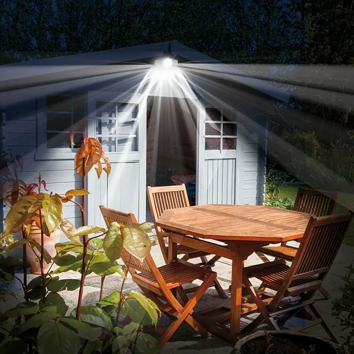 Proyector LED solar Beyong Bight X3 - Blanco frío, 1.000 lm - Comedor al aire libre con mesa y sillas de madera, iluminado por un brillante foco en un cobertizo.
