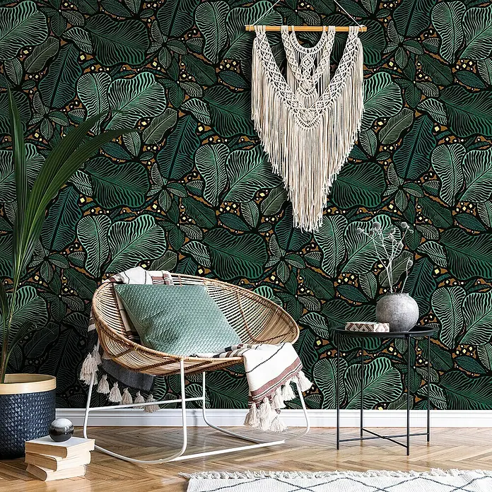 Papel pintado Trópico Verde - Trópico verde - Una habitación acogedora con una silla de ratán, plantas verdes y una decoración de pared de macramé frente a una pared estampada con hojas.