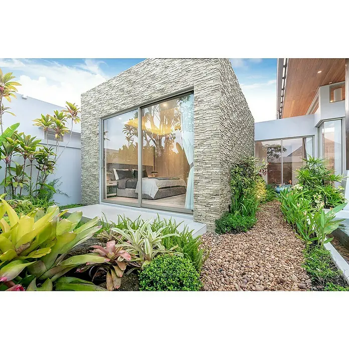 Stegu Verblendstein Kenya - 12 x 53,3 cm, Hellgrau, Steinoptik - Ein modernes Haus mit Steinverkleidung und großen Glasfenstern, umgeben von üppigem Grün. Der Fokus liegt auf Architektur und Gartendesign.