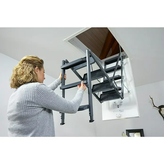 Sogem Escalera escamoteable Isoclic Pro Confort - 120 x 60 cm, Aislamiento térmico: 0,49 W/(m²K) - Una mujer baja una escalera de ático gris de una trampilla blanca en el techo, facilitando el acceso al ático.