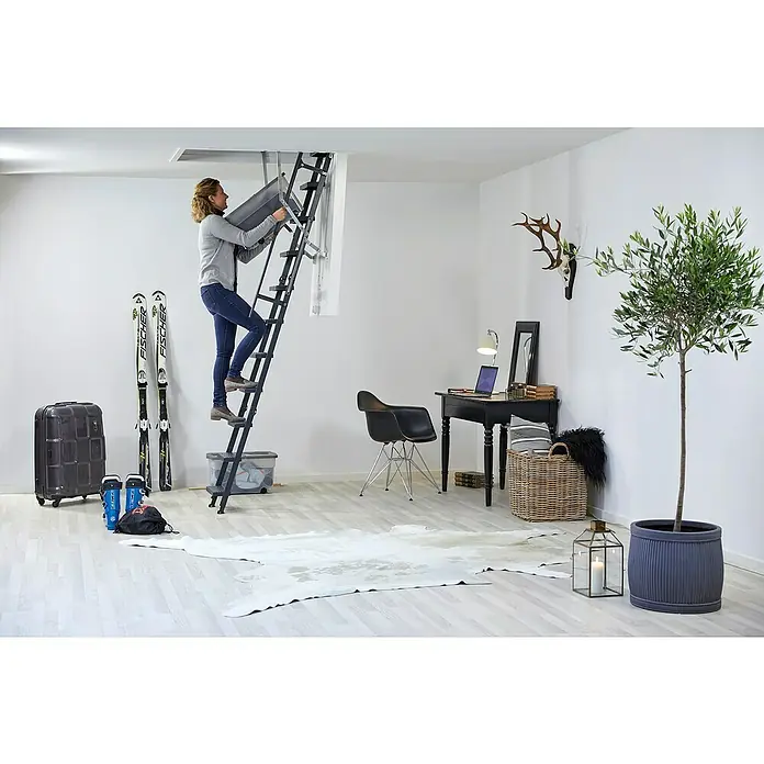 Sogem Escalera escamoteable Isoclic Pro Confort - 120 x 60 cm, Aislamiento térmico: 0,49 W/(m²K) - Una mujer sube por una escalera plegable de ático con una caja integrada en una habitación luminosa. La escalera es robusta y ahorra espacio.