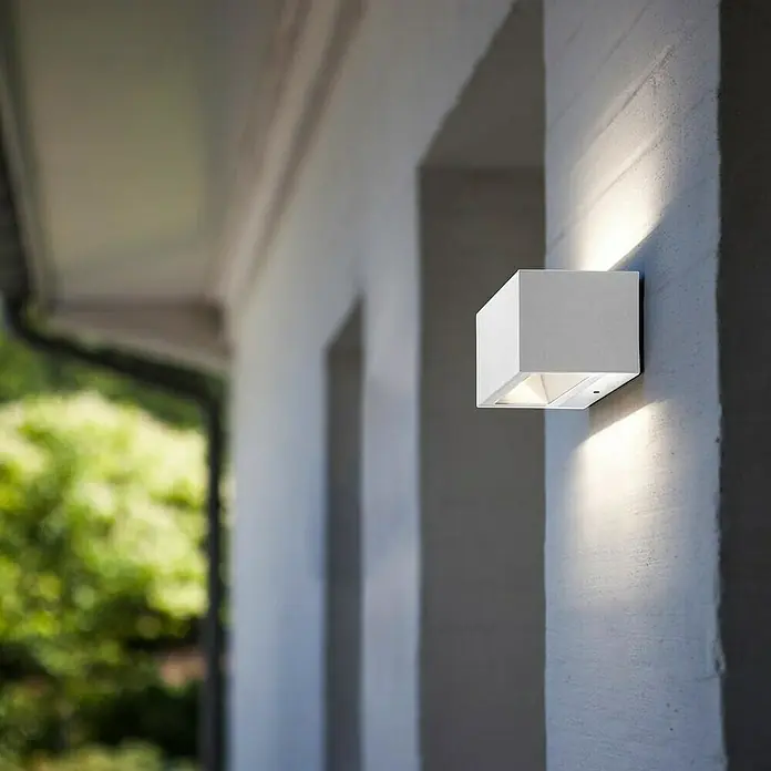 Lutec Aplique exterior LED Gemini - 10,5 W, 14 x 9 x 6,5 cm, Blanco, IP54 - Primer plano de una moderna lámpara de pared en forma de cubo, que emite una luz cálida y resalta la fachada arquitectónica.
