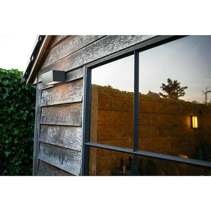Lutec LED-Außenwandleuchte Gemini - 10,5 W, 14 x 9 x 6,5 cm, Anthrazit, IP54 - Moderne Außenwandleuchte an einer Holzwand neben einem Fenster mit Spiegelung, die eine warme und einladende Atmosphäre schafft.