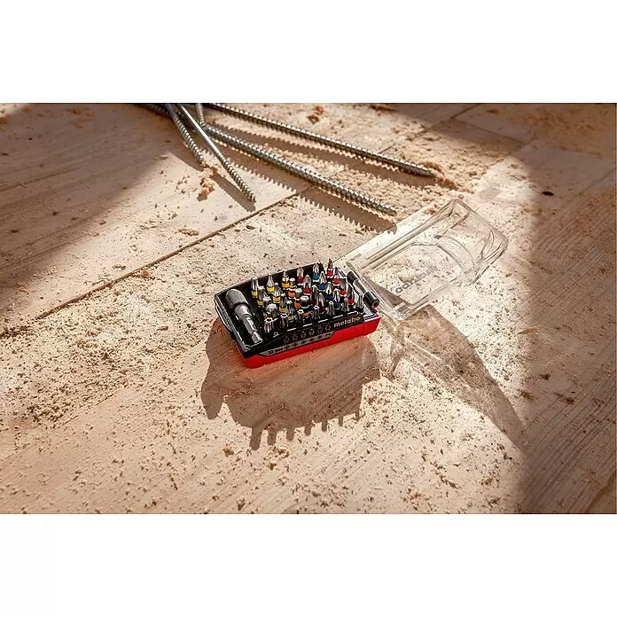 Metabo Bit-Box Akku Bit-Box - 32 -tlg. - Ein offenes rot-schwarzes Metabo Bit-Set mit Schraubendreher-Bits auf einer sägemehlbedeckten Holzoberfläche, neben langen Schrauben.