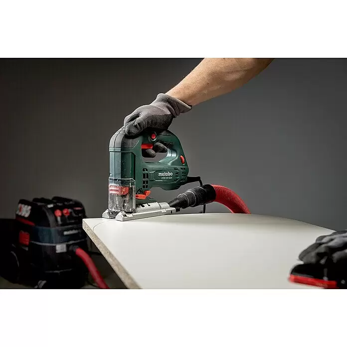 Metabo Elektro-Pendelhubstichsäge STEB 100 Quick - 1.000 /min - 3.100 /min, Max. Schnitttiefe in Holz: 100 mm, Anzahl Pendelhubstufen: 4 Stk. - Eine behandschuhte Hand bedient eine grüne Metabo Stichsäge, die an eine Staubabsaugung angeschlossen ist, und schneidet ein weißes Brett.