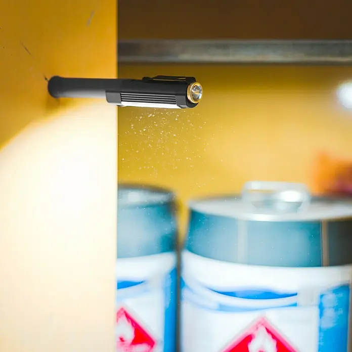 Nebo Tools Linterna Big Larry 2 - 3,2 x 2,5 x 19,7 cm, 40 lm - 500 lm - Primer plano de una luz de trabajo negra conectada a una superficie amarilla, que proyecta luz sobre dos latas borrosas.