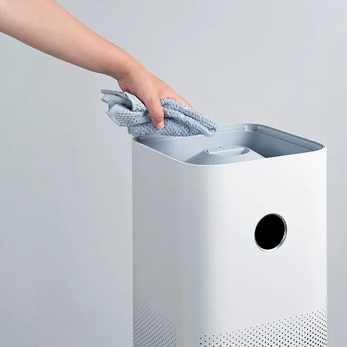 Xiaomi Luftreiniger Smart Air Purifier 4 Pro - L x B x H: 27,5 x 27,5 x 68 cm, Filtert bis zu 99,97 % aller Schadstoffe in einer Größe von 0,3 Micrometer - Das Bild zeigt einen Luftreiniger, der mit einem hellblauen Tuch gereinigt wird. Der Luftreiniger ist weiß und hat ein minimalistisches Design.