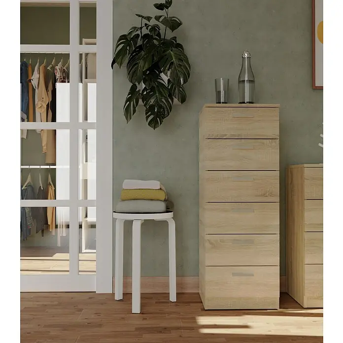 Muebles Pitarch Tibet Set muebles de dormitorio - Roble cambrian, 3 pzs. - Interior con una cómoda de madera clara con cajones, un taburete blanco con ropa y una planta en la pared.