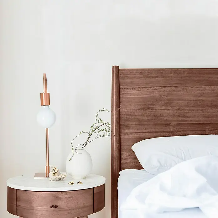 La Pajarita Pátina - Nogal, 75 ml, Mate - Primer plano de un dormitorio con una mesita de noche de mármol y cobre, una lámpara y una cama de madera con ropa de cama blanca.