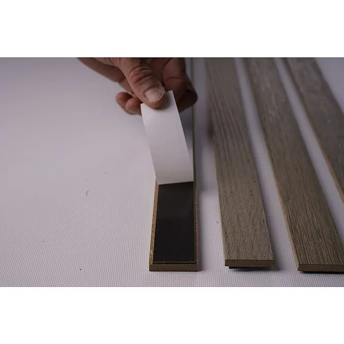 Panel de pared decorativo Sticks - Roble gris, 250 cm x 3,45 cm x 9 mm, 4 ud. - Una mano tira de la película protectora de una tira adhesiva en un suelo laminado, junto a otros listones laminados.