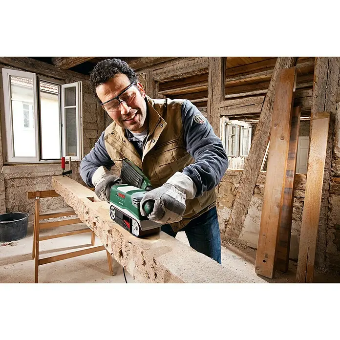 Bosch Elektro-Bandschleifmaschine PBS 75 AE Set - 750 W - Ein Mann benutzt einen Bosch Bandschleifer, um ein Holzstück zu schleifen. Der Bandschleifer ist grün und schwarz und scheint effizient zu sein.