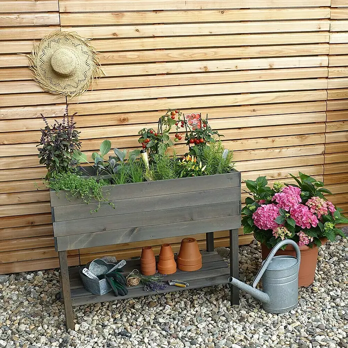 Promadino Kräuterhochbeet - L x B x H: 100 x 50 x 80 cm, Grau - Bild eines erhöhten grauen Pflanzgefäßes aus Holz mit verschiedenen Pflanzen, Gartengeräten und einer Gießkanne vor einer Holzwand.