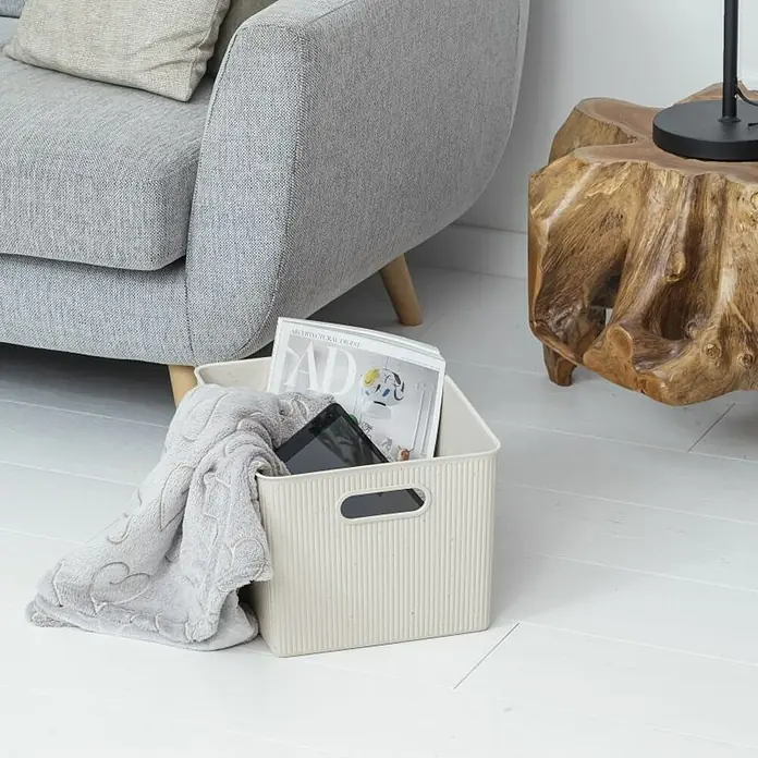 Tatay Baobab Cesta para la ropa - 29,4 x 38,5 x 23 cm, Plástico, Beige - Una cesta de almacenamiento beige en un piso de madera, llena con una manta, una revista y una tableta, se encuentra al lado de un sillón gris y una mesa de madera.