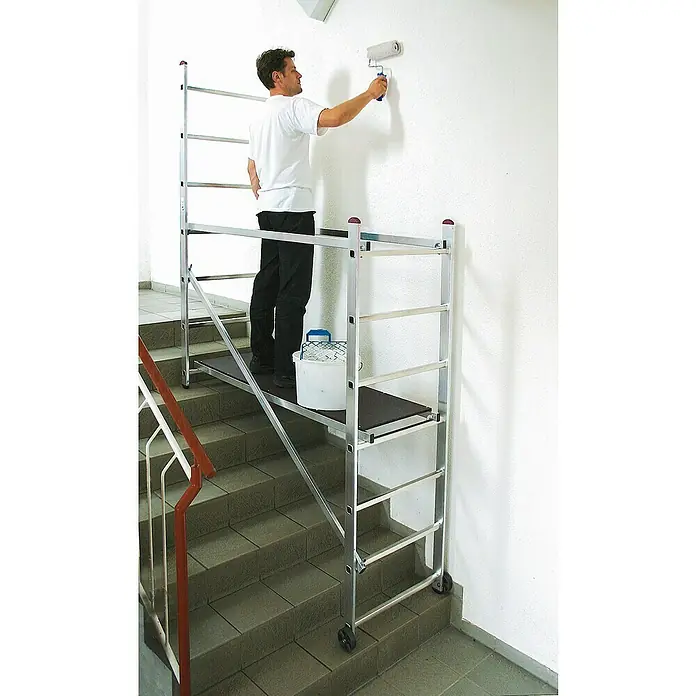 Krause Corda Kleingerüst - Arbeitshöhe: 3 m - Ein Mann streicht eine Wand mit einer Farbwalze, stehend auf einem Gerüst auf einer Treppe. Das Gerüst ist aus Aluminium und hat Rollen.