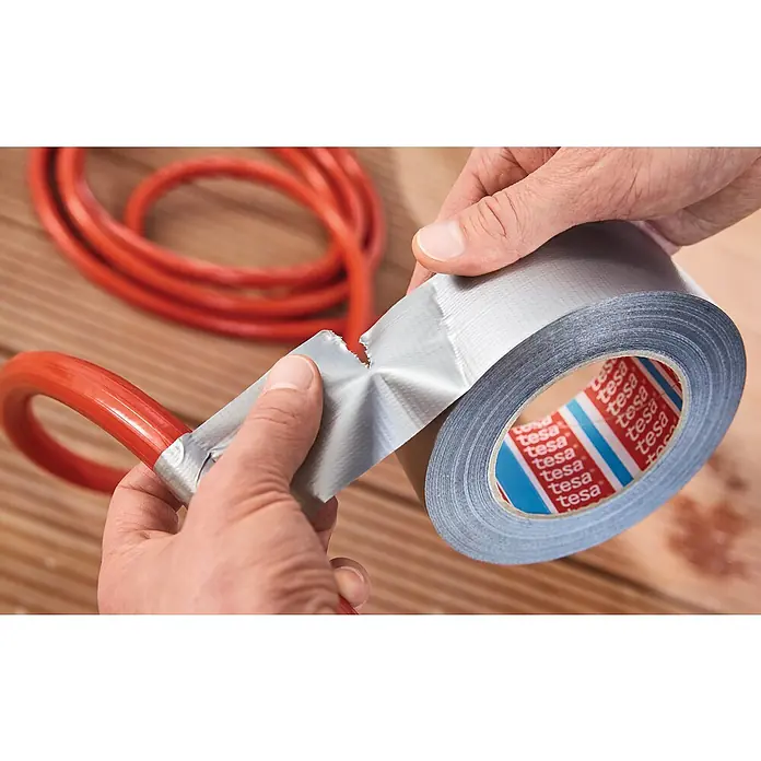 Tesa Extra Power Folienband Universal - Silber, 25 m x 50 mm - Eine Rolle graues Tesa-Gewebeband wird verwendet, um ein rotes Kabel zu reparieren. Das Klebeband ist teilweise abgerissen.