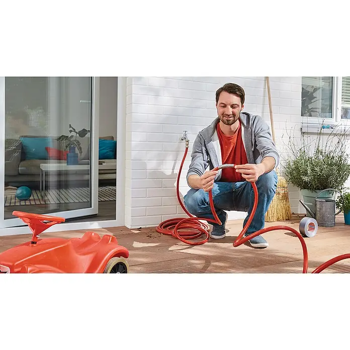 Tesa Extra Power Folienband Universal - Silber, 25 m x 50 mm - Mann schließt einen roten Gartenschlauch an einen Wasserhahn im Freien an, während er auf einer Holzterrasse kniet.