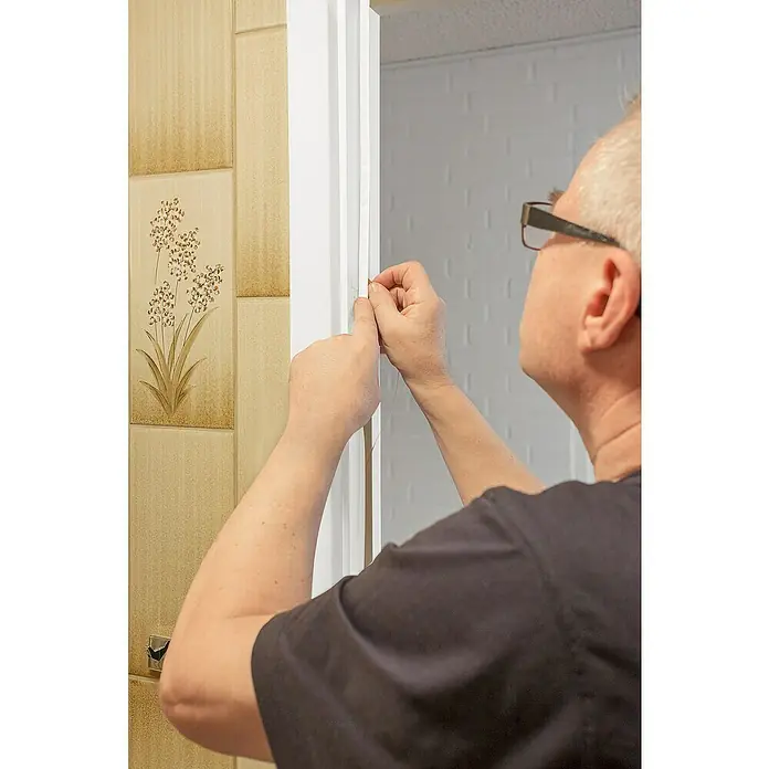 Primo Universaldichtung Densa 25 - Weiß, 6 m, Passend für: Fenster, Türen, Möbel mit Spaltenbreite 2 – 5 mm - Ein Mann befestigt sorgfältig eine weiße Zierleiste an einer Tür mit einer dünnen Schnur, um sie zu fixieren.