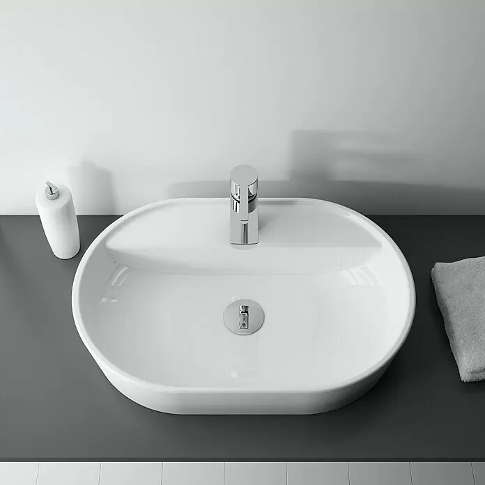 Inspira Aufsatzwaschbecken - 62 x 41 cm, Keramik, Weiß - Weißes ovales Keramikwaschbecken mit Chromarmatur auf einer dunkelgrauen Arbeitsplatte. Daneben ein weißer Seifenspender und ein graues Handtuch.